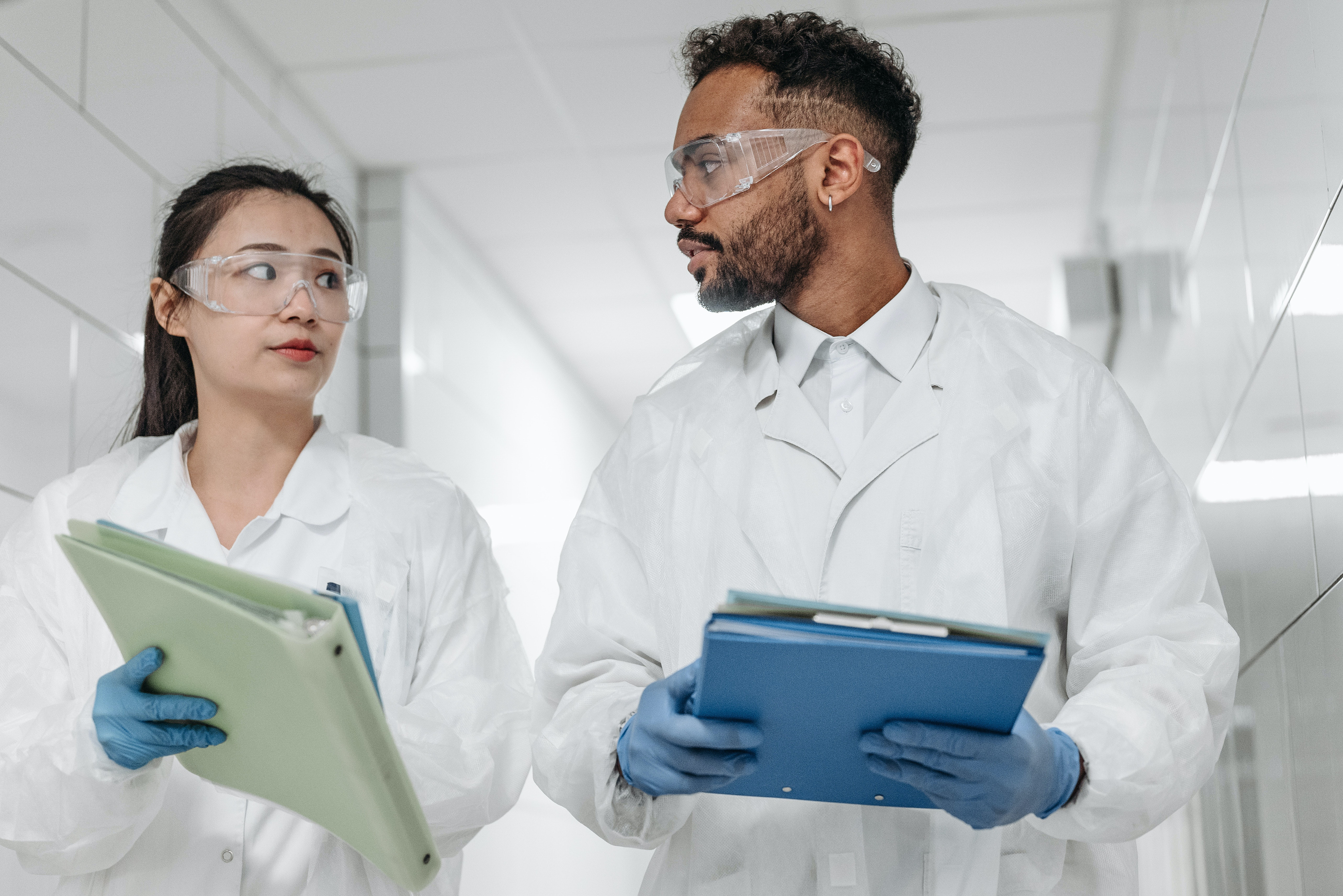 Two health care workers wearing glasses and lab coats walk down a hallway with clipboards in discussion