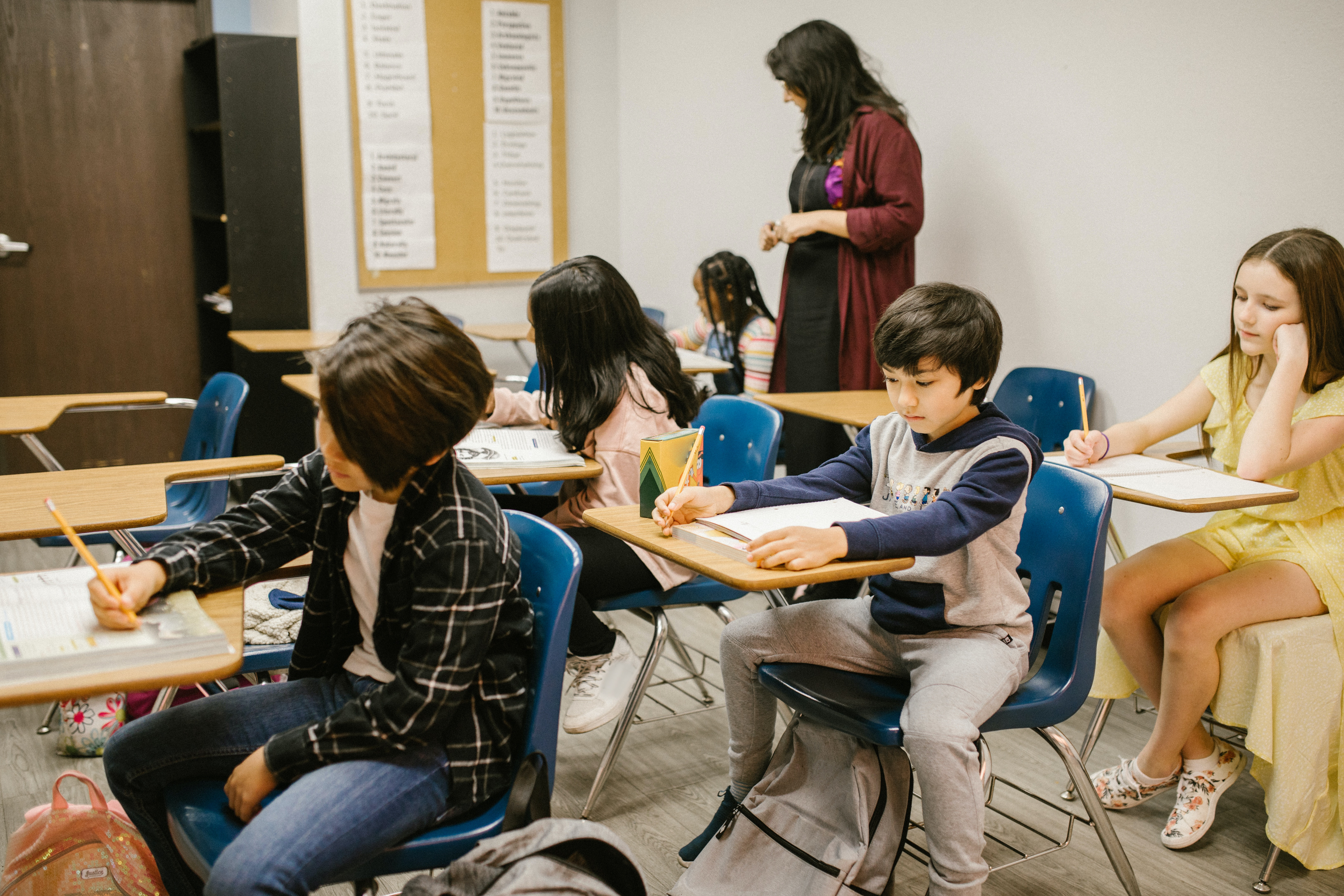 Children sitting at desks in a classroom working on schoolwork with a teacher standing over them in the back of the room