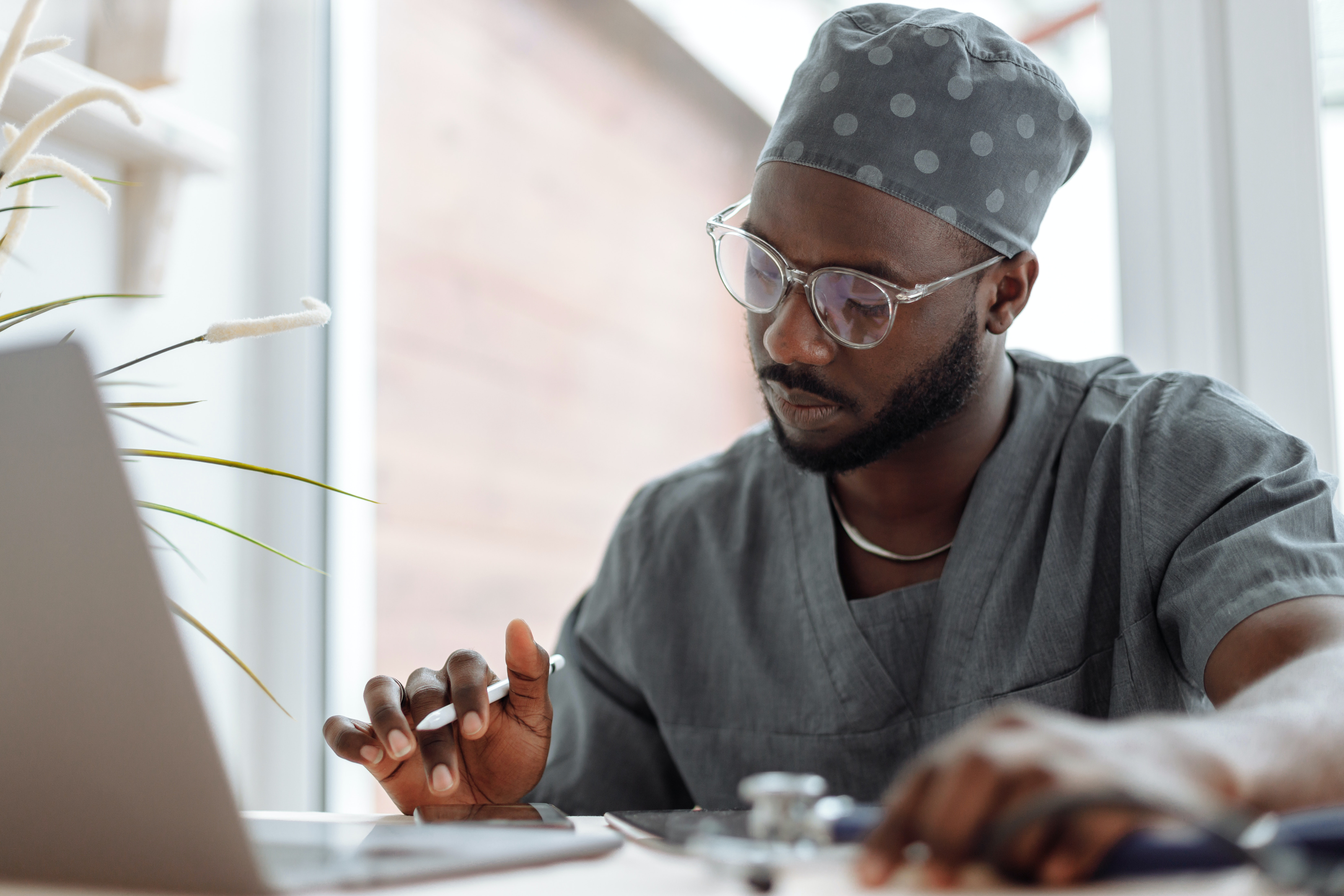 A medical student sits at a table dressed in scrubs with a computer, phone, stylus and stethoscope