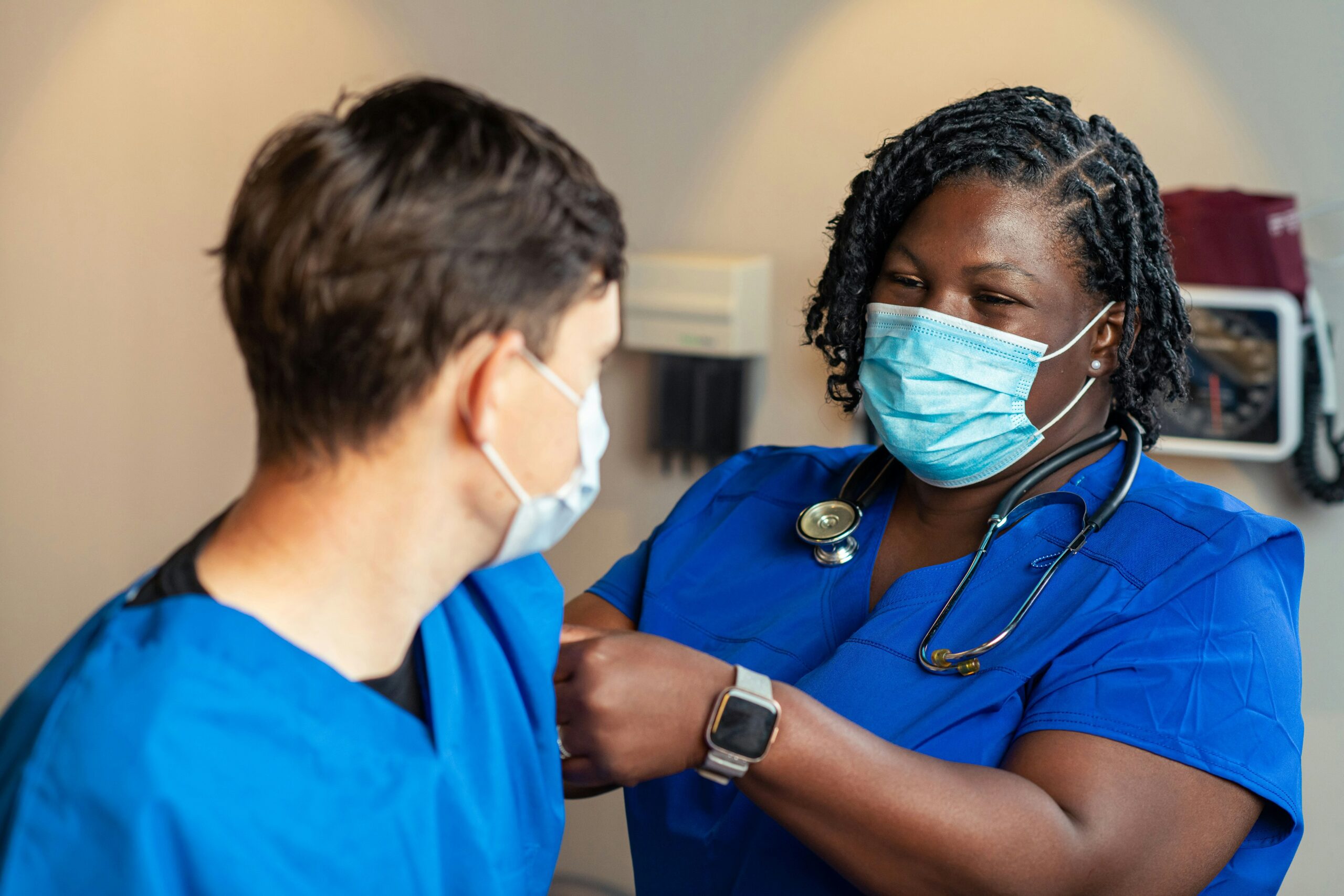 Two health professionals wearing masks and blue scrubs, one is looking at and taking the blood pressure of the other