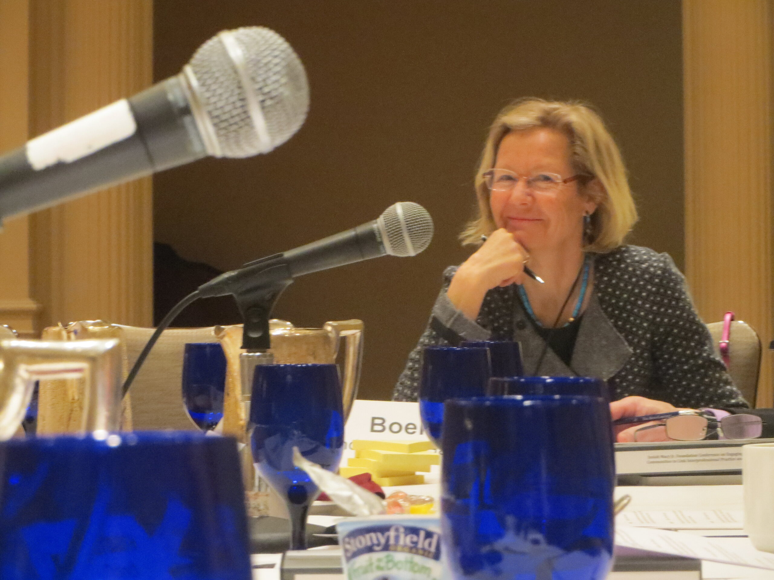 A photo of microphones and conference materials on a table with a woman smiling in the background
