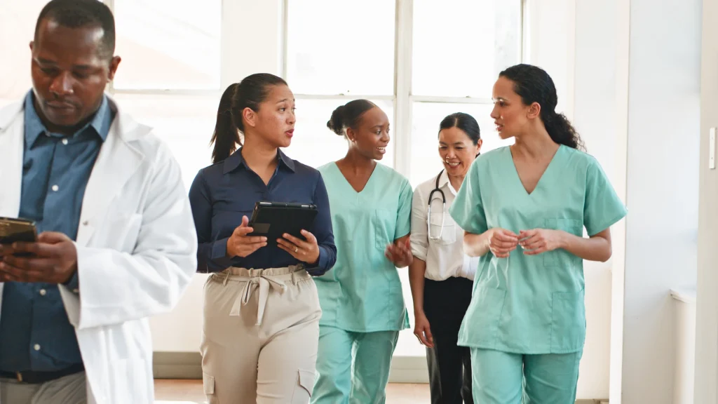 Healthcare workers in scrubs and office attire walking together, interacting in a bright medical hallway.